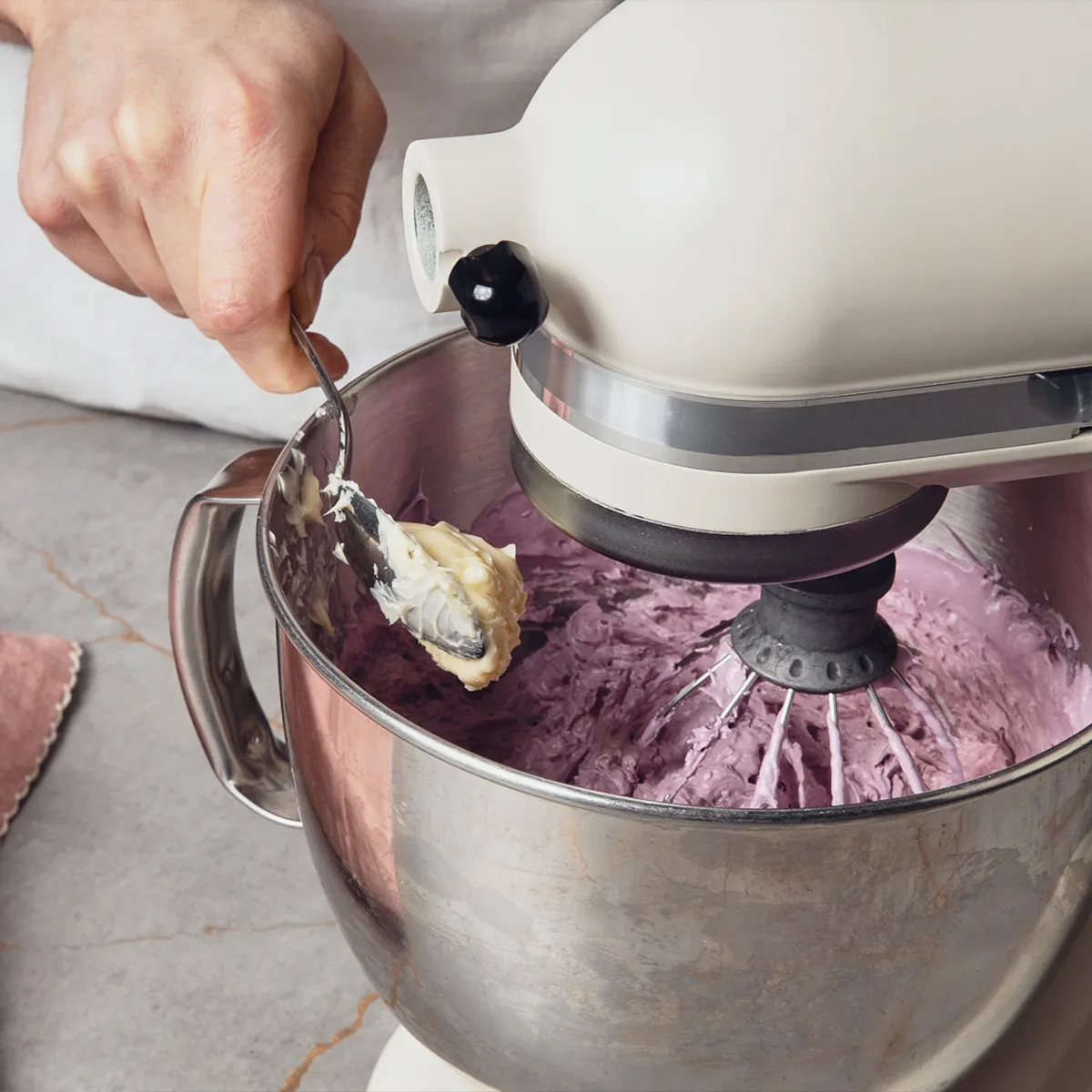A hand adds a spoon of butter to the bowl of a stand mixer which is filled with a lilac cream.
