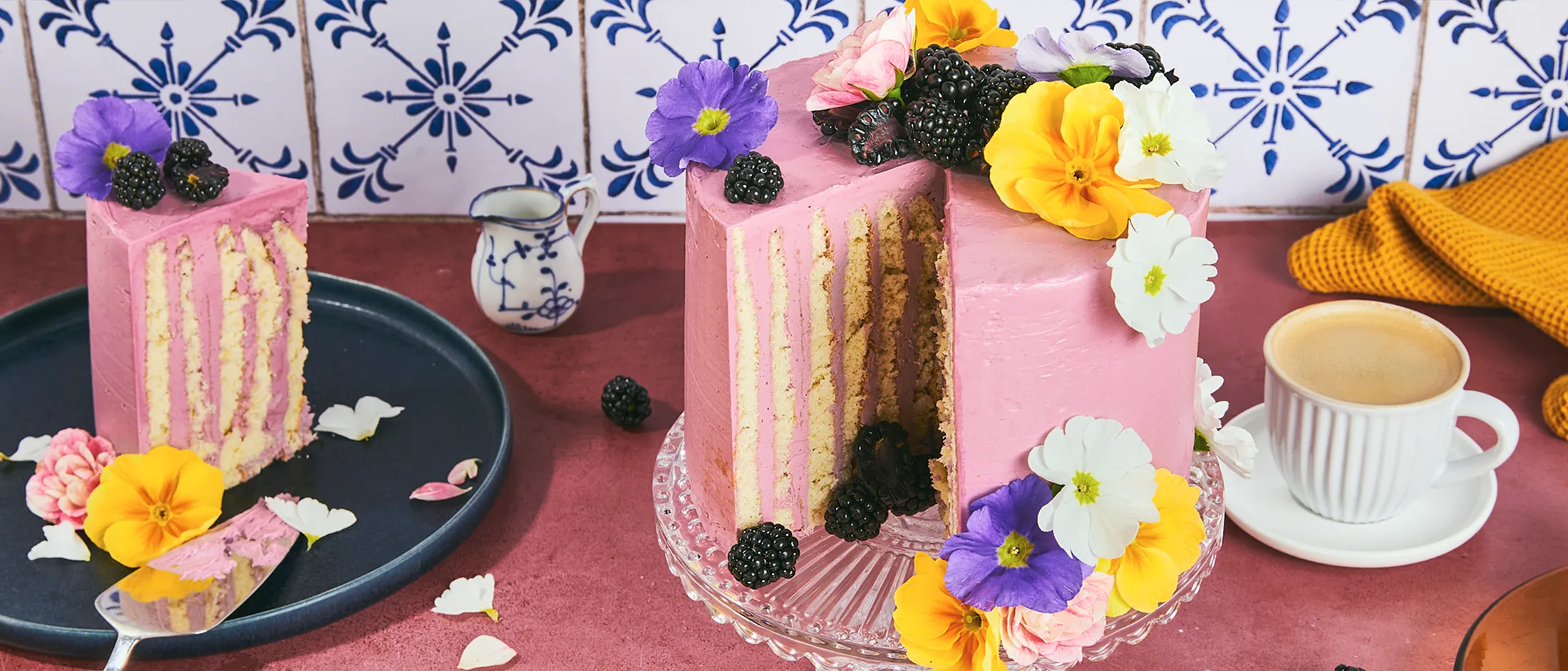 Una torta con righe verticali alle more, con una fetta tagliata, su un'alzatina in vetro appoggiata su un tavolo rosa, circondata da altre stoviglie.