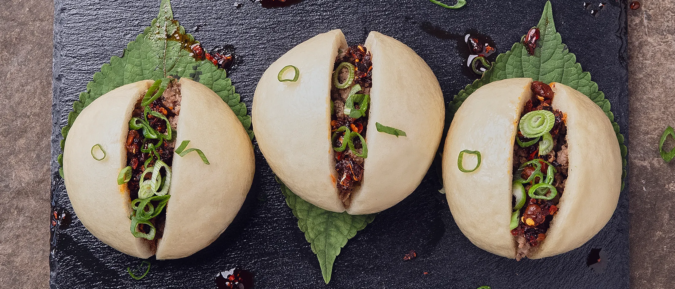 Drei gedämpfte, mit Schweinefleisch gefüllte Teigtaschen, aufgeschnitten und mit gewürztem Hackfleisch, in Scheiben geschnittenen Frühlingszwiebeln und Chili Crisp gefüllt, angerichtet auf Shiso-Blättern auf einer schwarzen Schieferplatte.