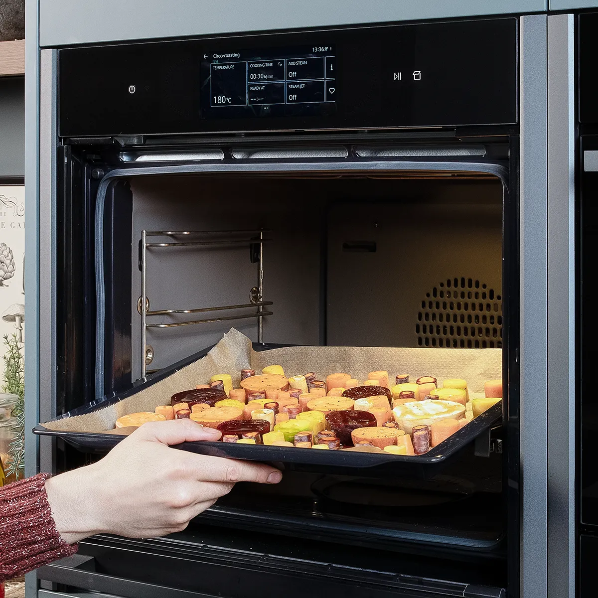 Ein Backblech mit farbenfrohem Wurzelgemüse wird in einen geöffneten Backofen geschoben.
