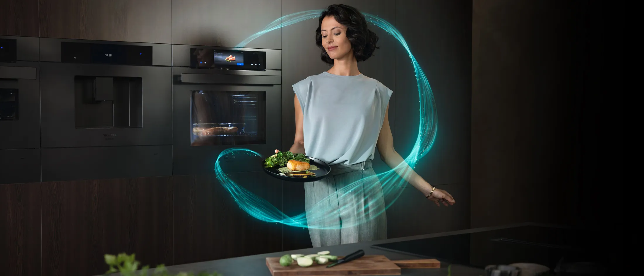 Moderne keuken met de matzwarte keukenapparaten van Siemens en in een blauwgrijze outfit geklede dame met een maaltijd vers uit de oven.