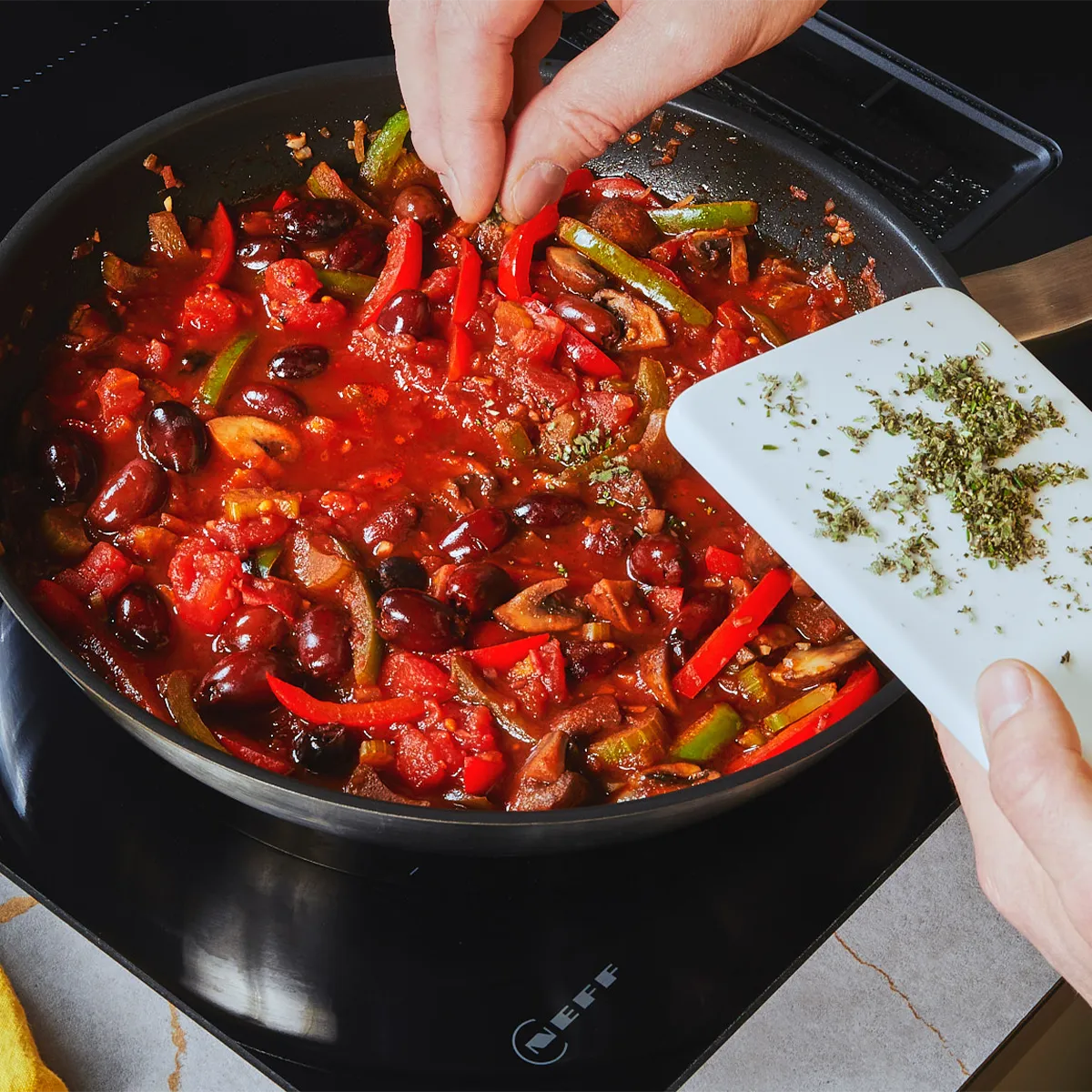 Hände streuen getrocknete Kräuter in eine Pfanne mit Tomaten-, Paprika- und Oliveneintopf auf einem Neff-Induktionskochfeld; kleines Schneidebrett und gelbes Tuch in der Nähe.