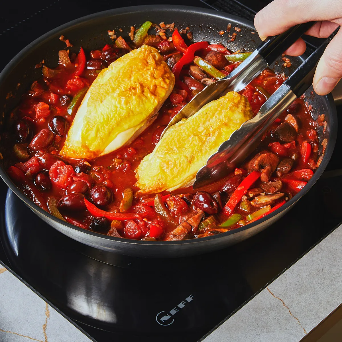 Hand mit Zange legt zwei goldbraune Hähnchenbrüste in eine Tomaten-, Paprika- und Olivenpfanne auf einem Neff-Induktionskochfeld; gelbes Tuch auf der Arbeitsplatte.