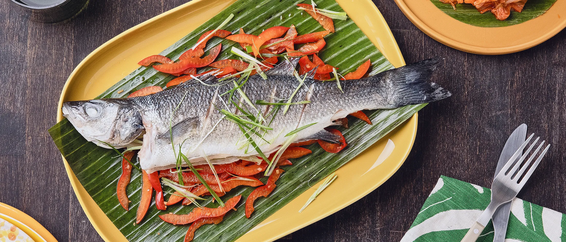 Ein ganzer gedämpfter Wolfsbarsch, angerichtet auf einem Bananenblatt mit in Scheiben geschnittenen roten Paprikaschoten und Frühlingszwiebeln, präsentiert auf einem gelben Teller mit Kimchi als Beilage.