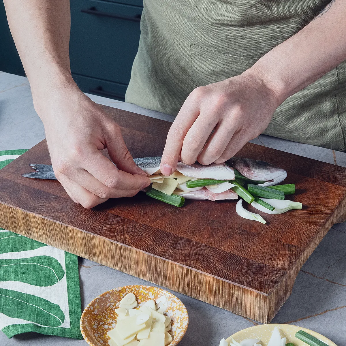 Person stuffing a whole raw sea bass with sliced ginger and scallions on a wooden cutting board.
