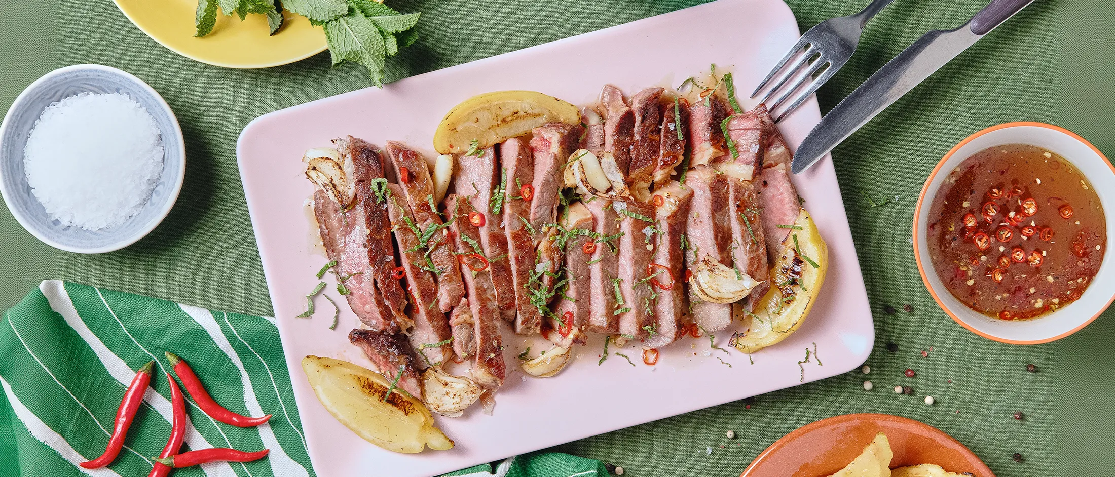 Ein gegrilltes und in Streifen geschnittenes Steak liegt auf einem rechteckigen, rosafarbenen Teller, der auf einer grünen Tischdecke steht, umgeben von Geschirr.