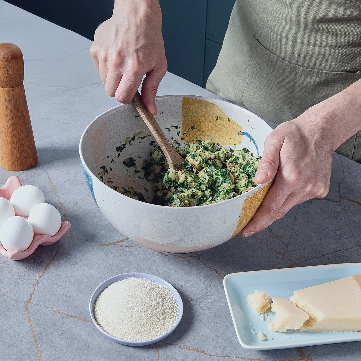 Person mixing chopped spinach, bread, and cheese in a bowl with a wooden spoon, surrounded by eggs, pepper mill, grated cheese, and breadcrumbs.
