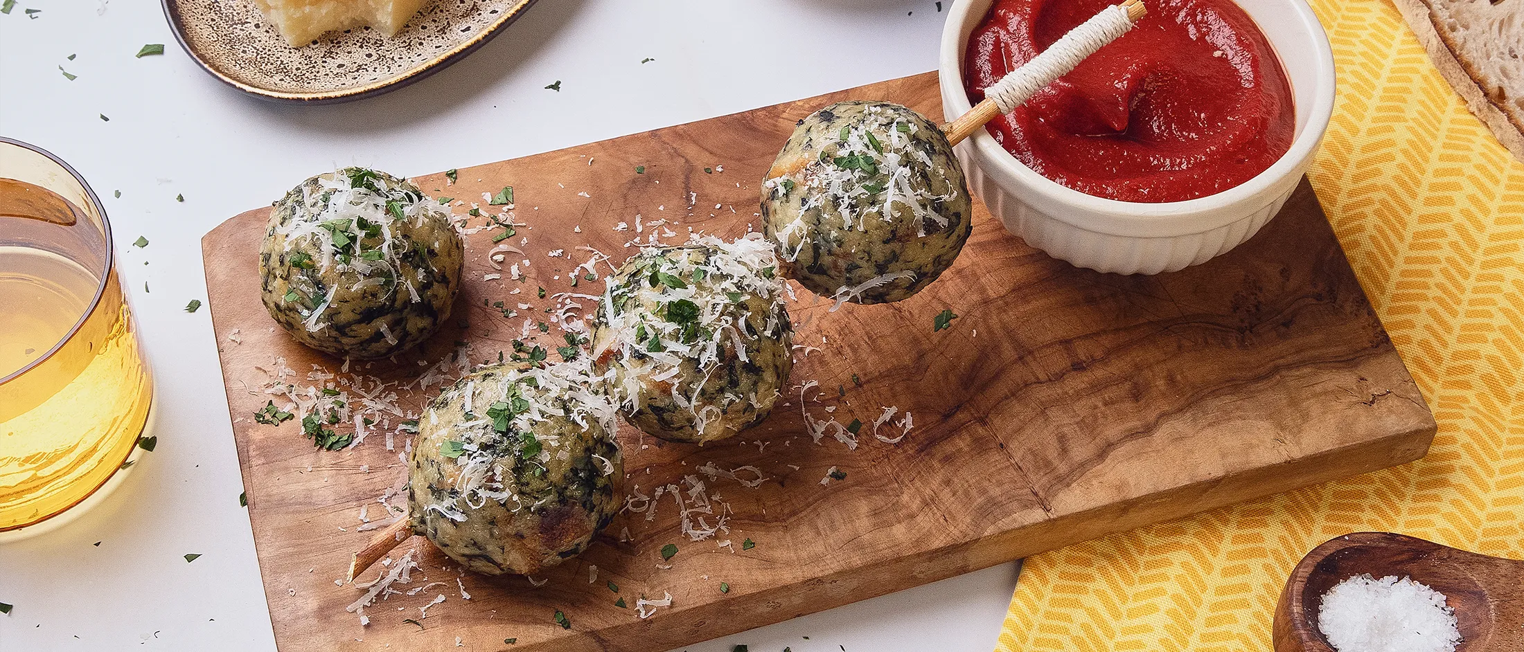 Vier Spinat- und Käseknödel auf einem Holzschneidebrett, mit geriebenem Käse und Petersilie bestreut und mit Tomaten-Dattel-Sauce in einer Schüssel sowie rustikalem Brot als Beilage serviert.