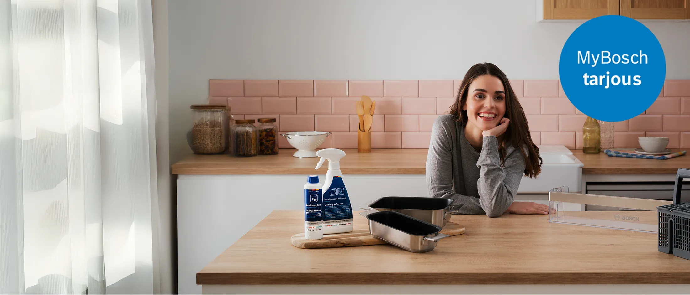 Hymyilevä nainen nojaa keittiön puiseen tasoon, jolla on esillä erilaisia Bosch-astianpesukoneen lisävarusteita ja puhdistusaineita. Kuvan yläkulmassa on sininen ympyrä, jossa lukee "MyBosch tarjous".
