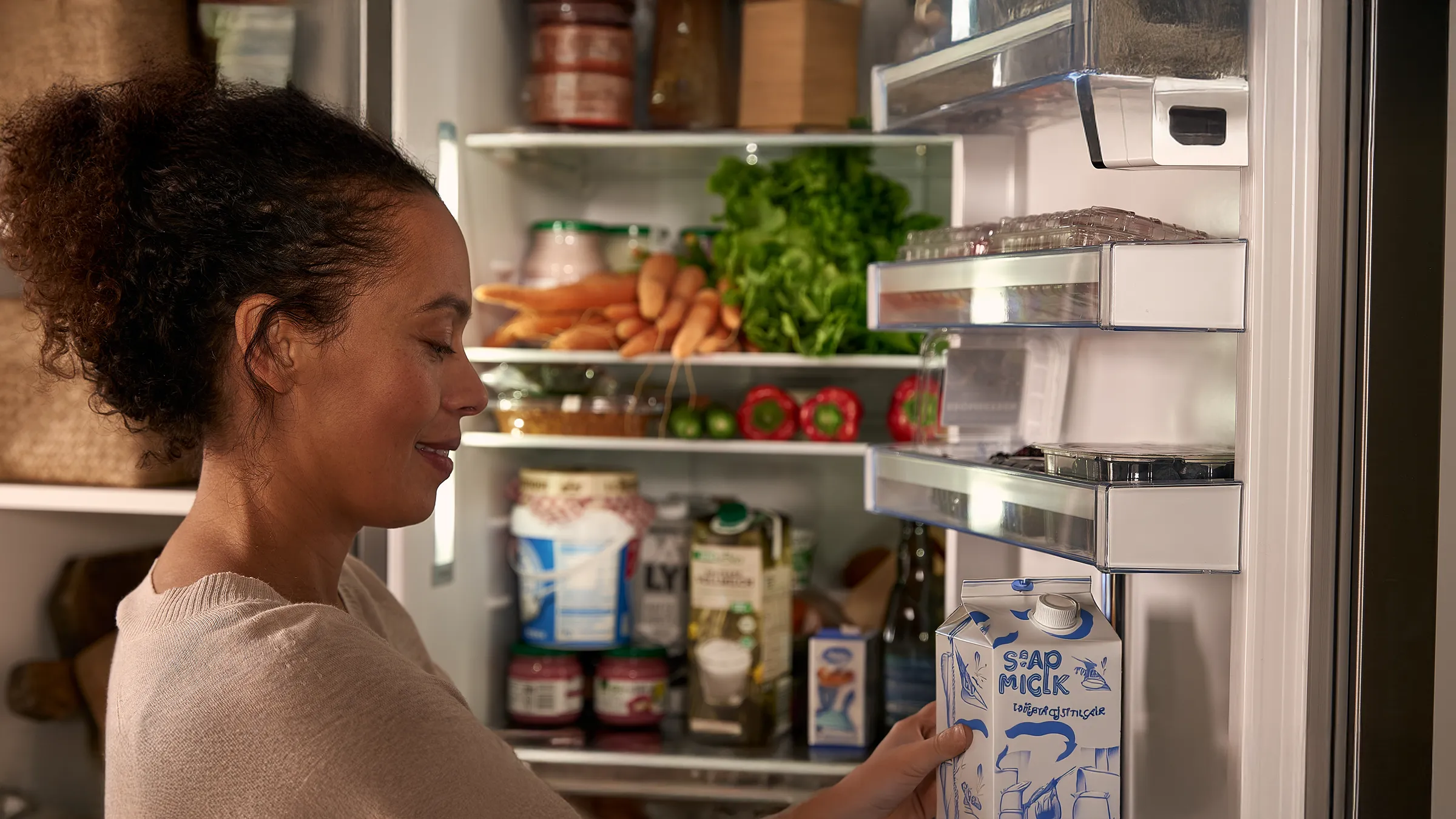 Person taking a carton of soy milk from an open refrigerator filled with vegetables, jars, and cartons.
