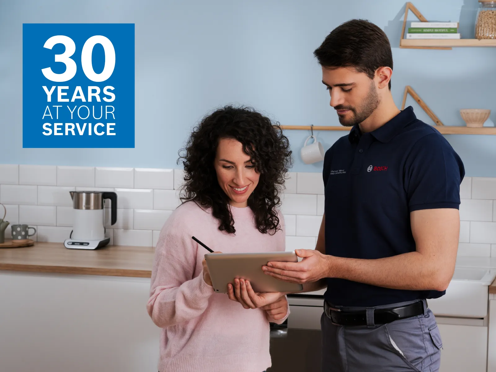 En Bosch-tekniker i handsker reparerer et apparat i et moderne køkken, mens en kvinde ser på. Værktøj ligger på bordet.