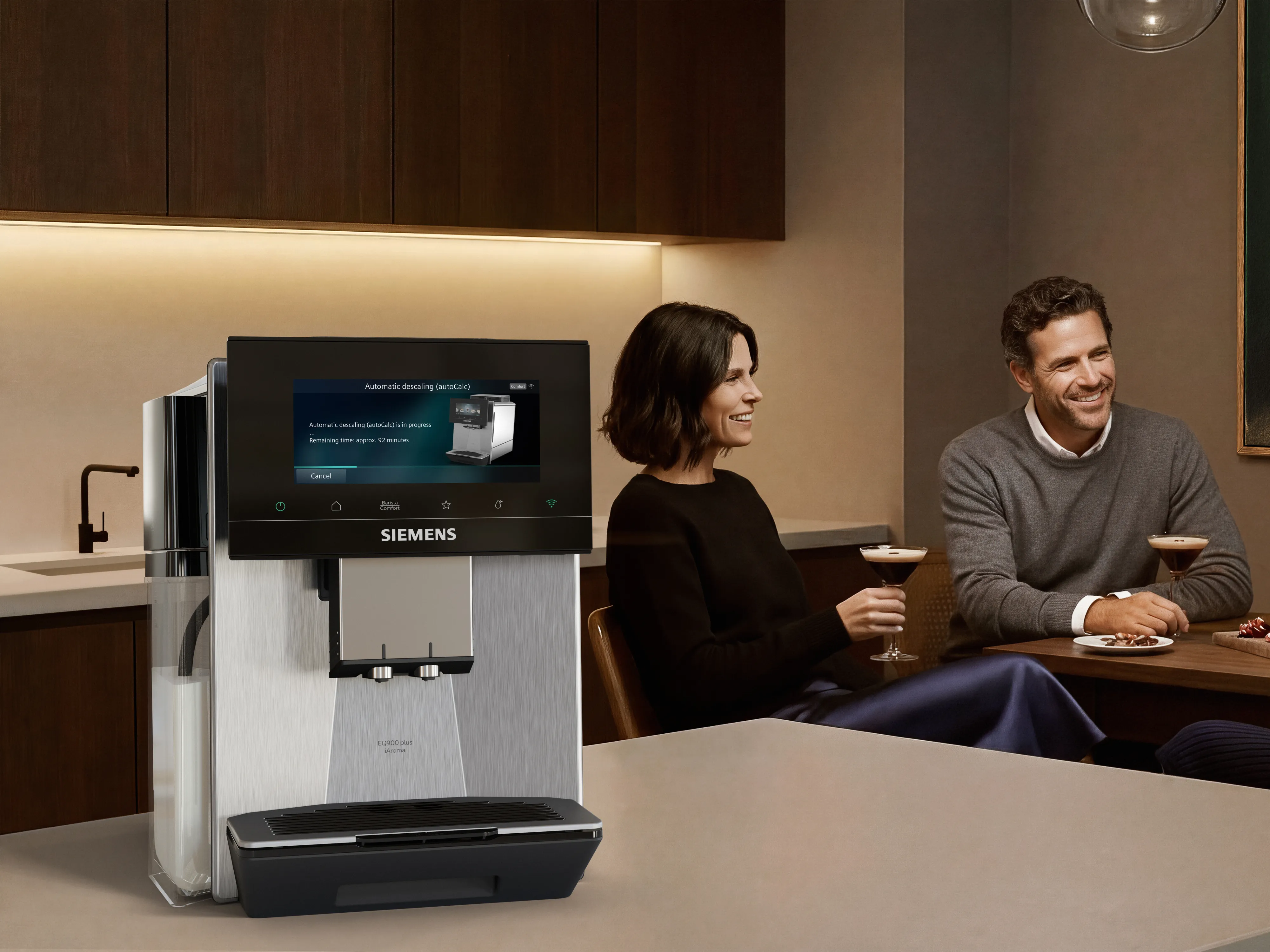 Siemens EQ900 plus iAroma on a modern kitchen counter showing an automatic descaling cycle, while two people sitting at a wooden table in the foreground, smiling over cocktails and snacks in a warm, softly lit setting.