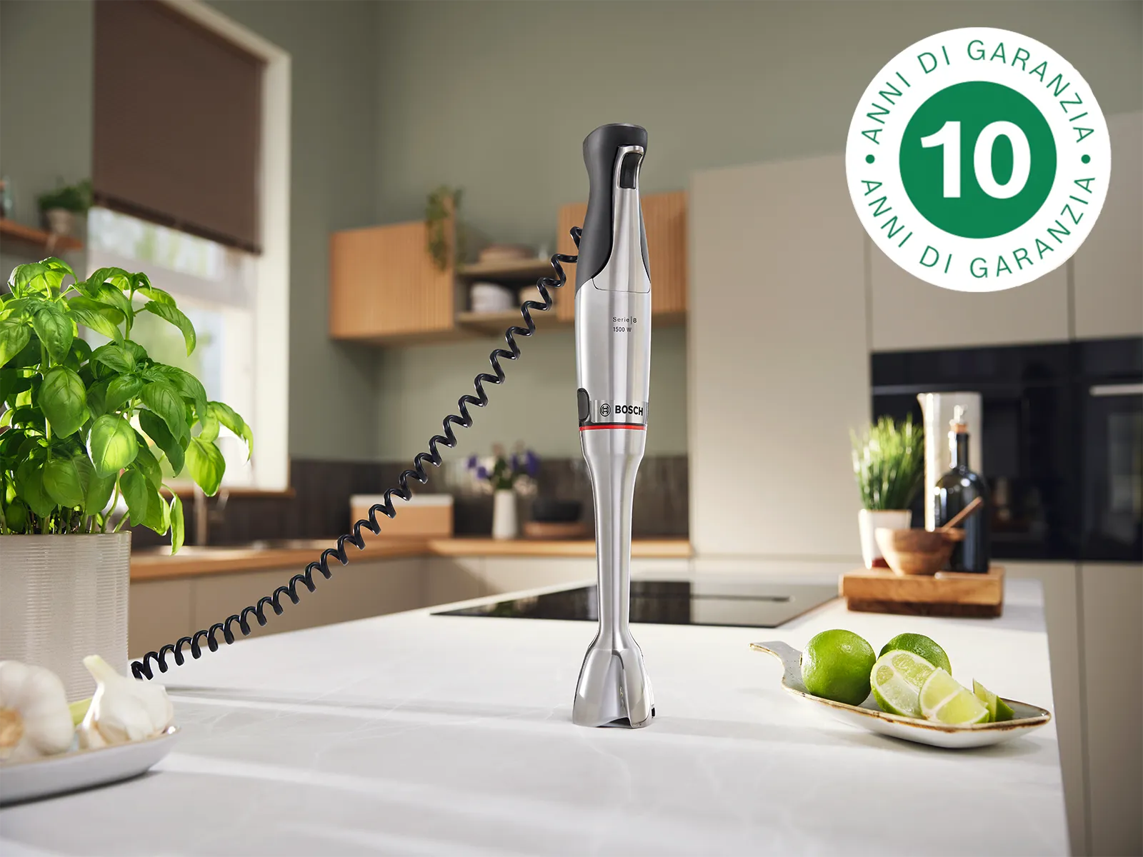 Frullatore a mano in piedi sul bancone della cucina con pianta di basilico, spicchi d'aglio e tagli di lime, in un ambiente cucina pulito e timbro con logo di garanzia del motore di 10 anni.