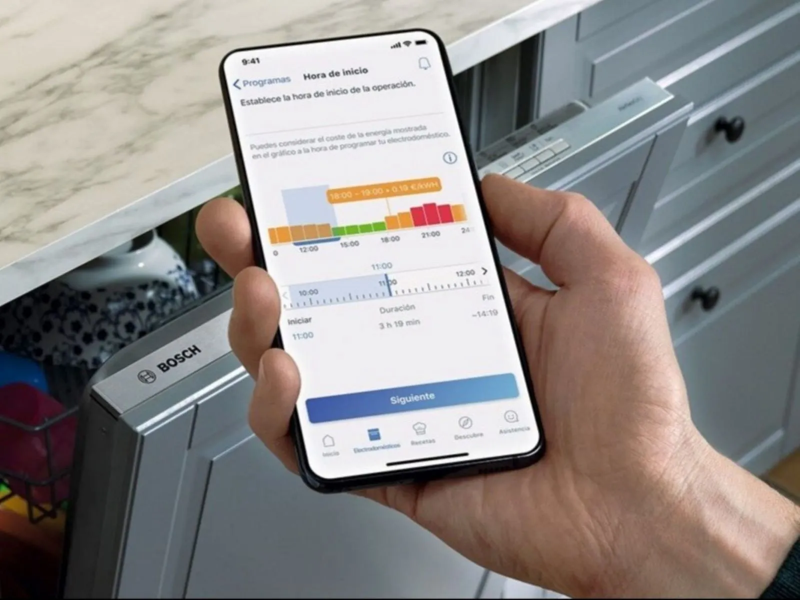 Una persona sostiene un teléfono inteligente que muestra una aplicación de programación para un lavavajillas Bosch, con configuraciones de uso de energía y tiempo visibles.