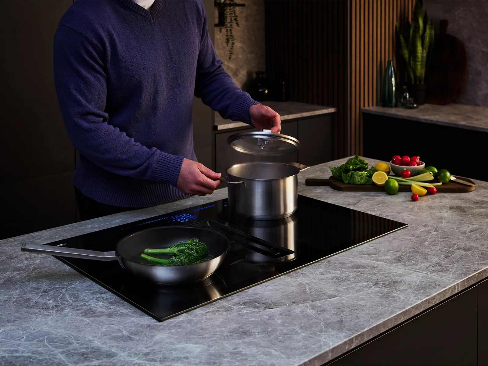 A man in a blue sweater cooks on a black Siemens induction cooktop, lifting a pot lid while broccoli simmers in a pan and fresh vegetables are prepped nearby.