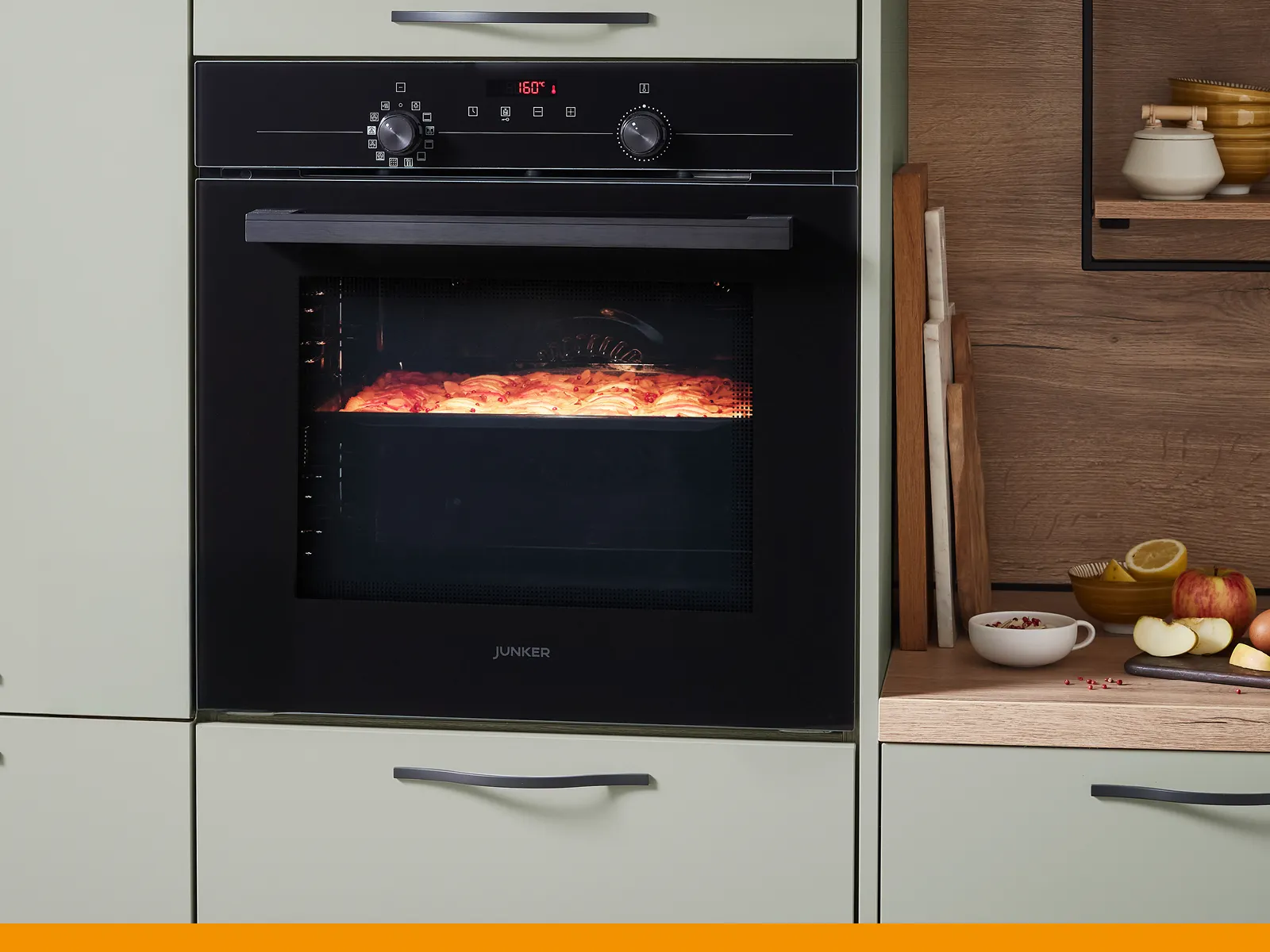 "A black Junker built-in oven in a sage green kitchen, with the interior light on showing a cake baking inside.