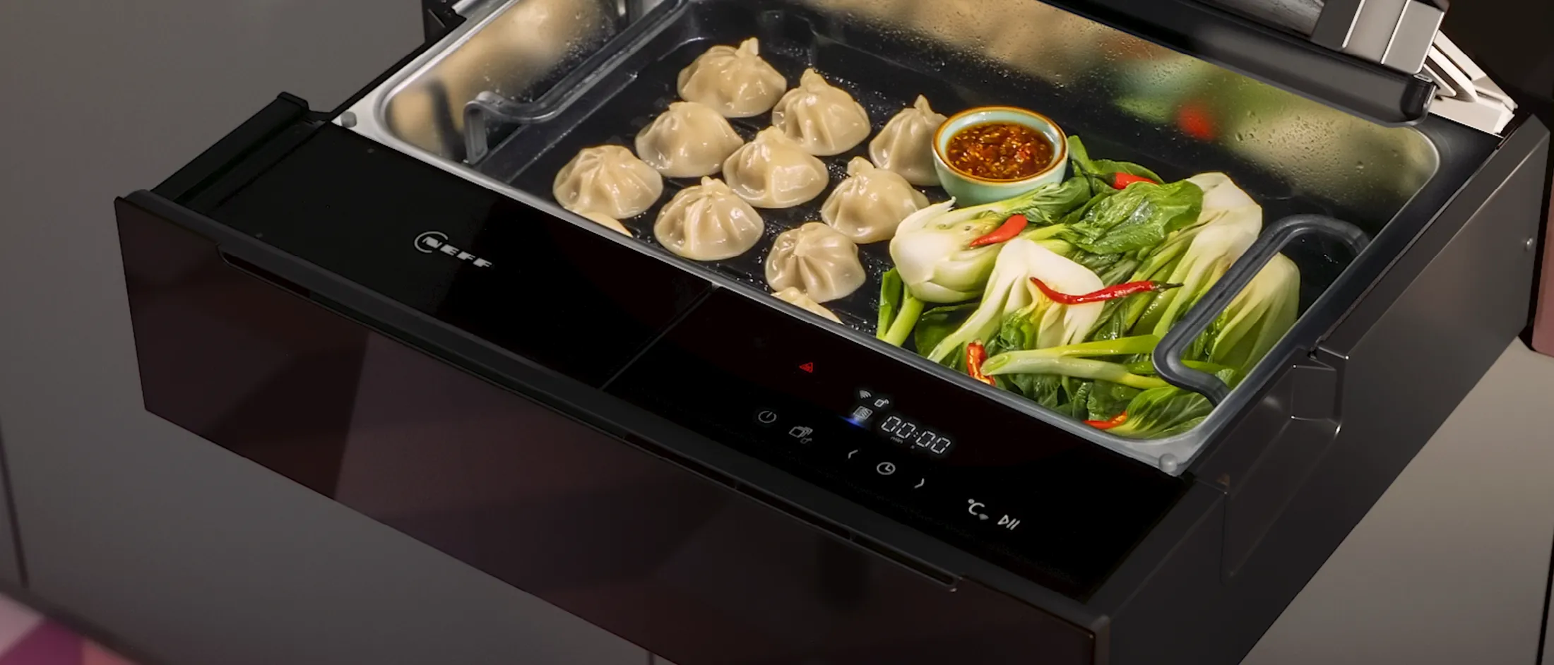 Open stoomlade van NEFF met negen gestoomde dumplings, kommetje chilisaus en gestoomde paksoi.