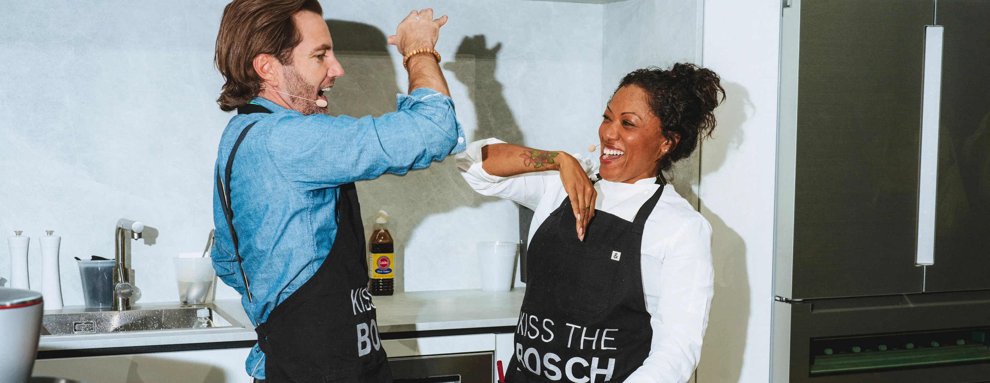 Two people wearing aprons in a kitchen, playfully interacting near a Bosch refrigerator.