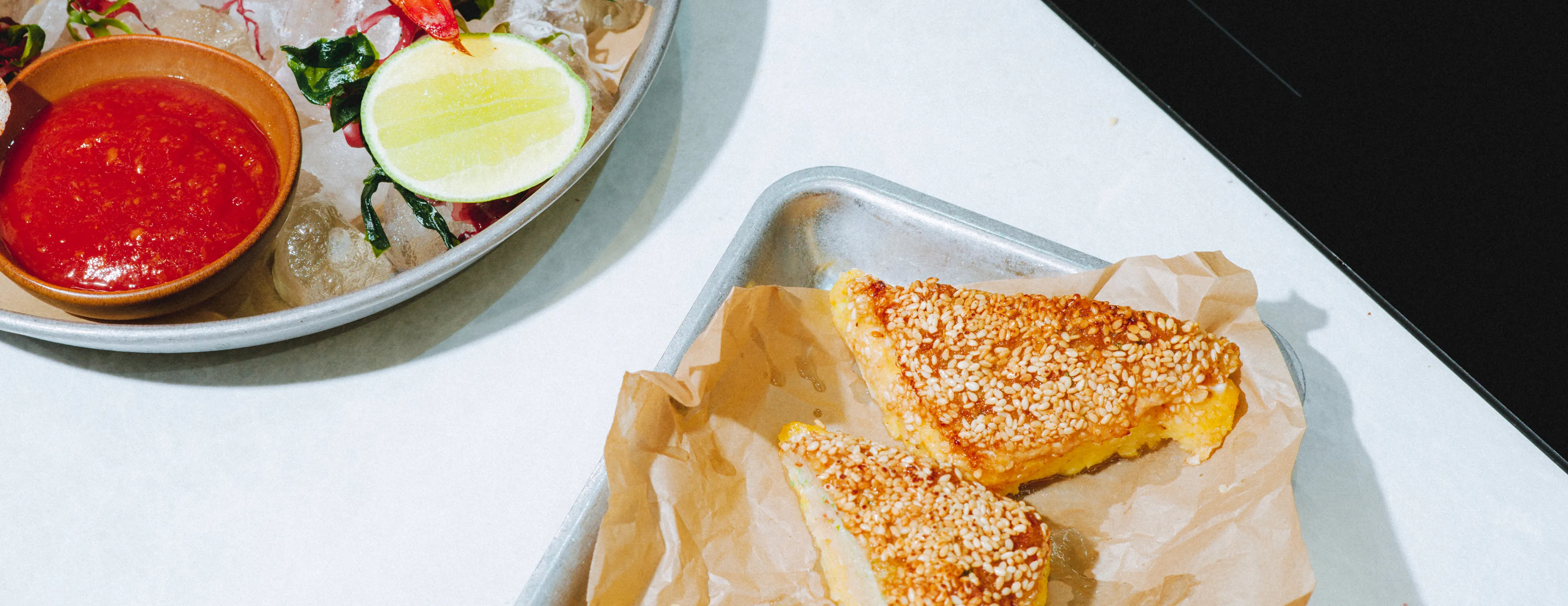 Two sesame-coated triangle toasts on a tray, with a bowl of red sauce and lime on the side.