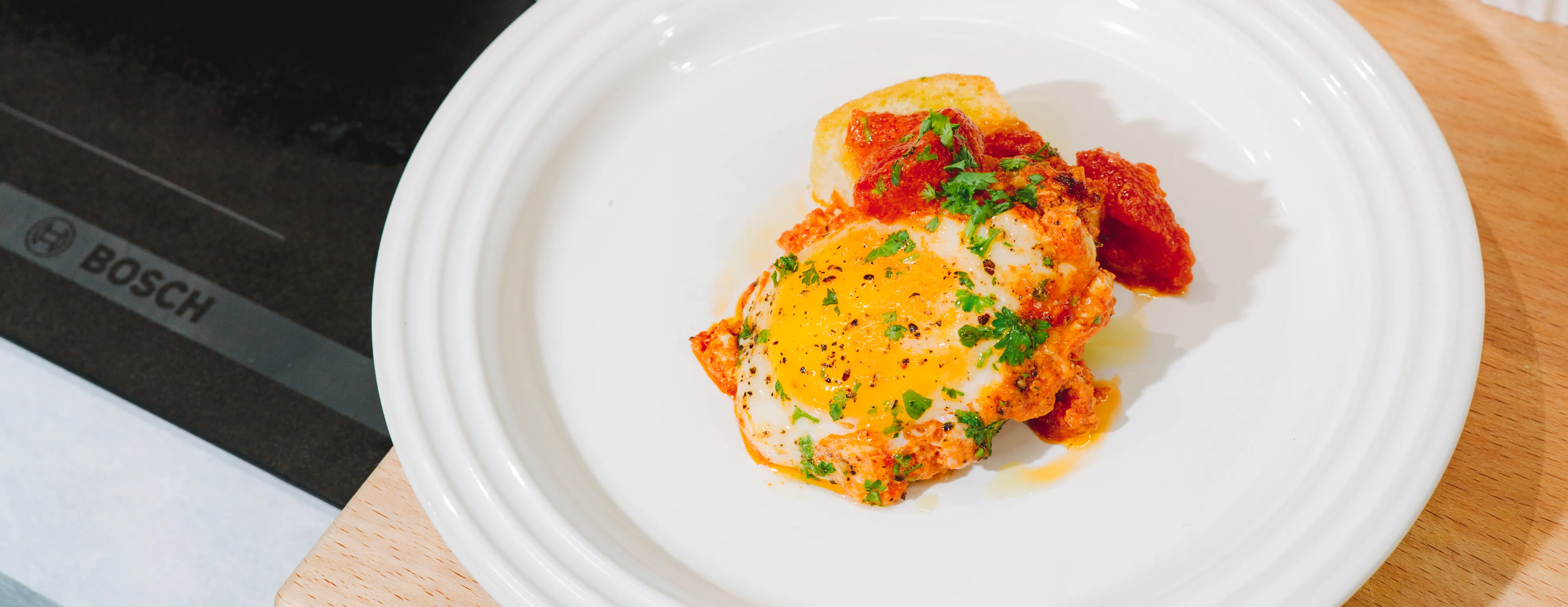 Fried egg with tomato sauce and herbs on white plate, Bosch logo visible on stovetop.
