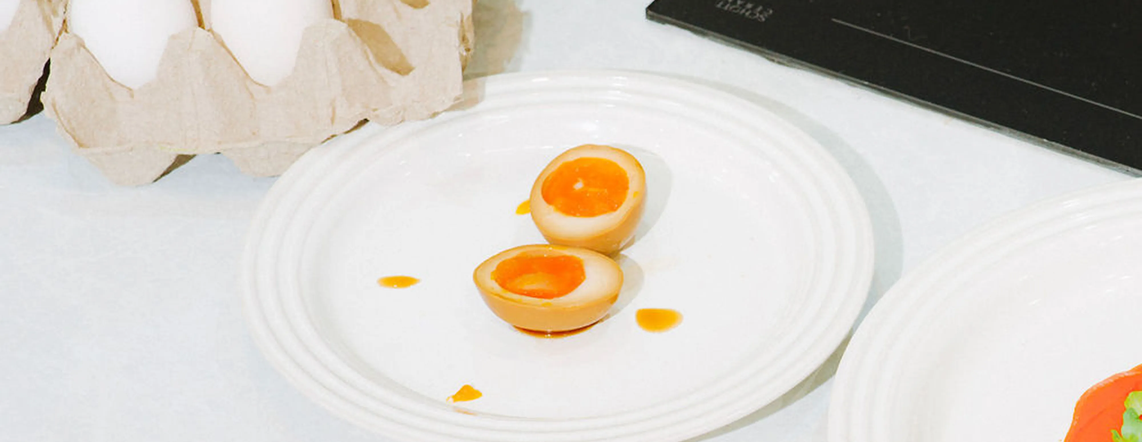 A halved soft-boiled egg on a white plate next to a carton of eggs on a kitchen countertop.