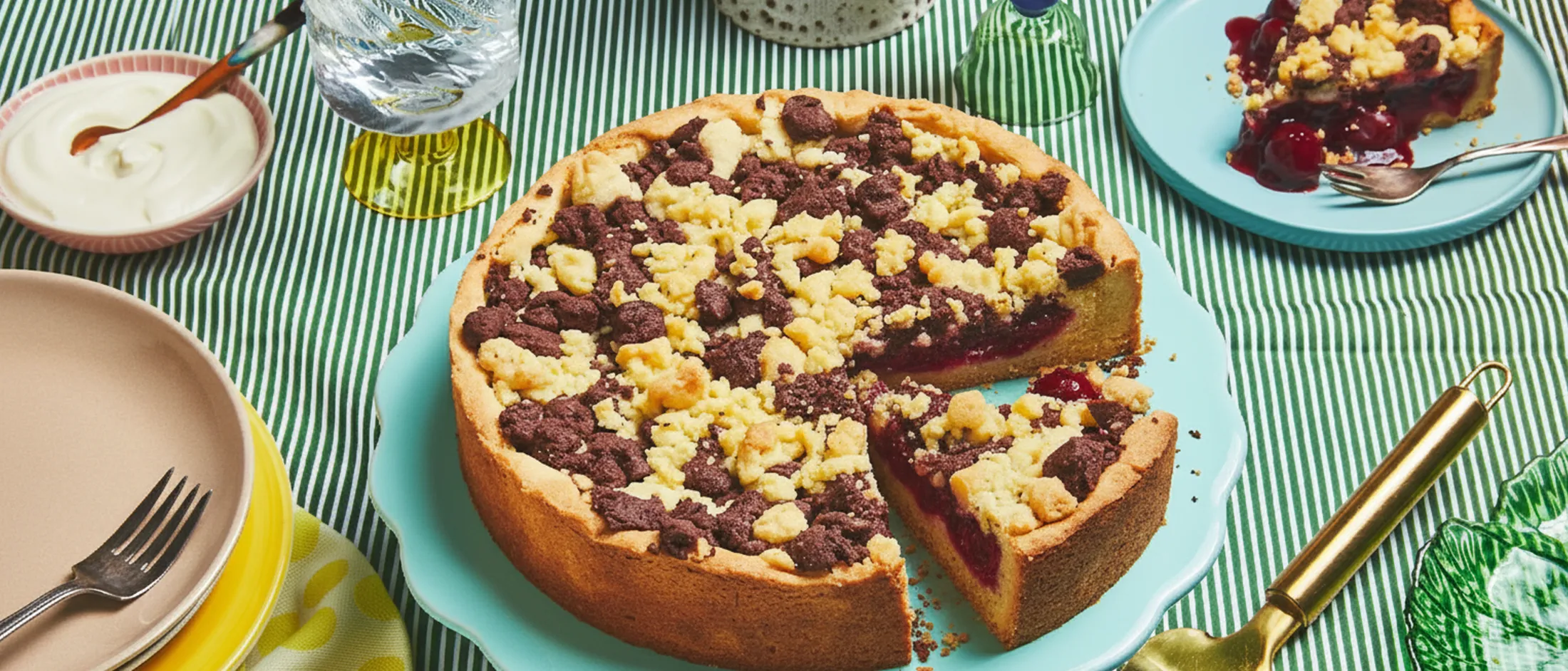 Ein Streuselkuchen mit Sauerkirschen auf einem hellblauen Tortenständer steht auf einer grünen Tischdecke, umgeben von anderem Geschirr.