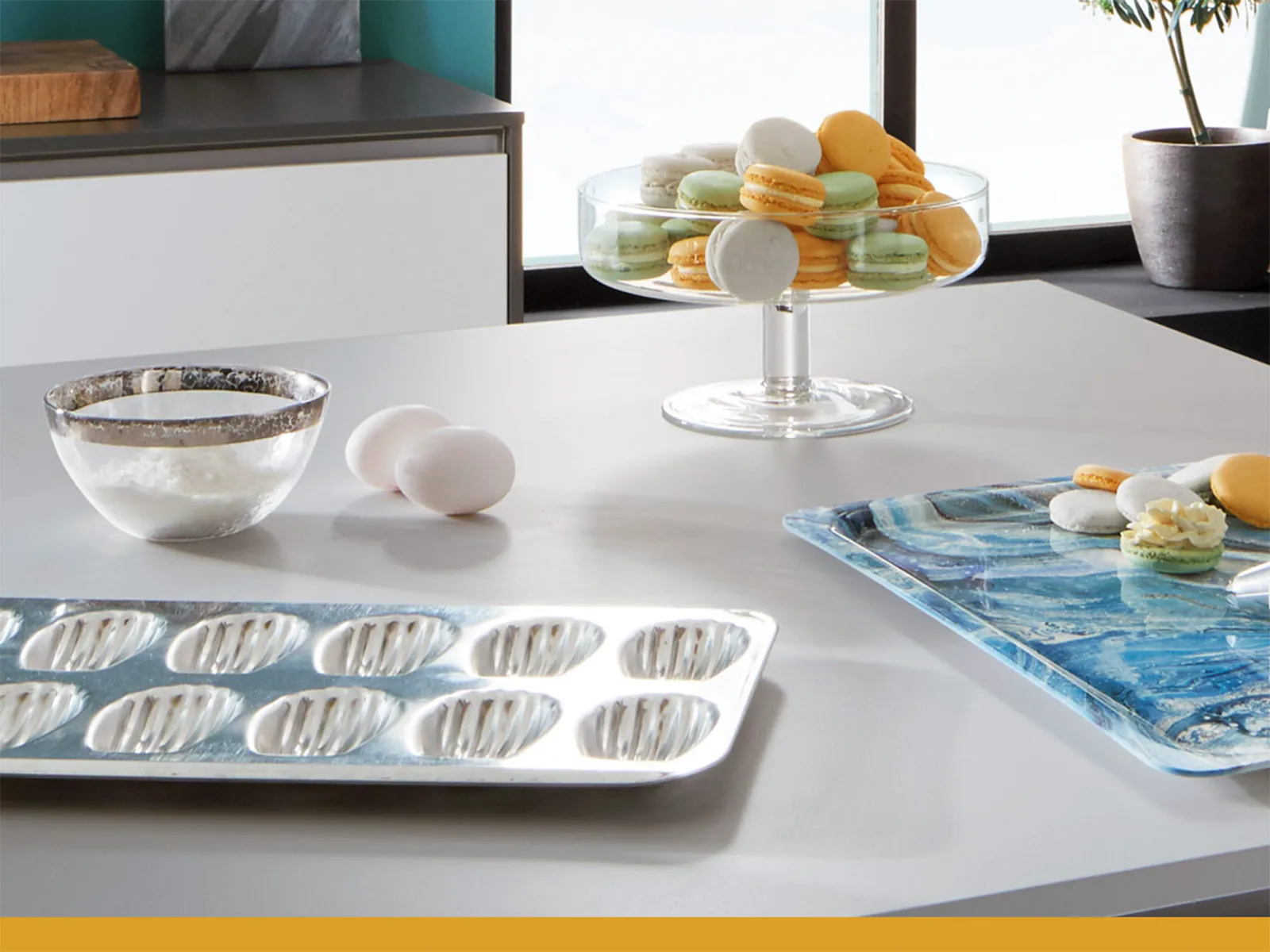 Arbeitsplatte mit Macaron-Formen, Zuckerdose, Eiern und einer Schale mit bunten Macarons. 