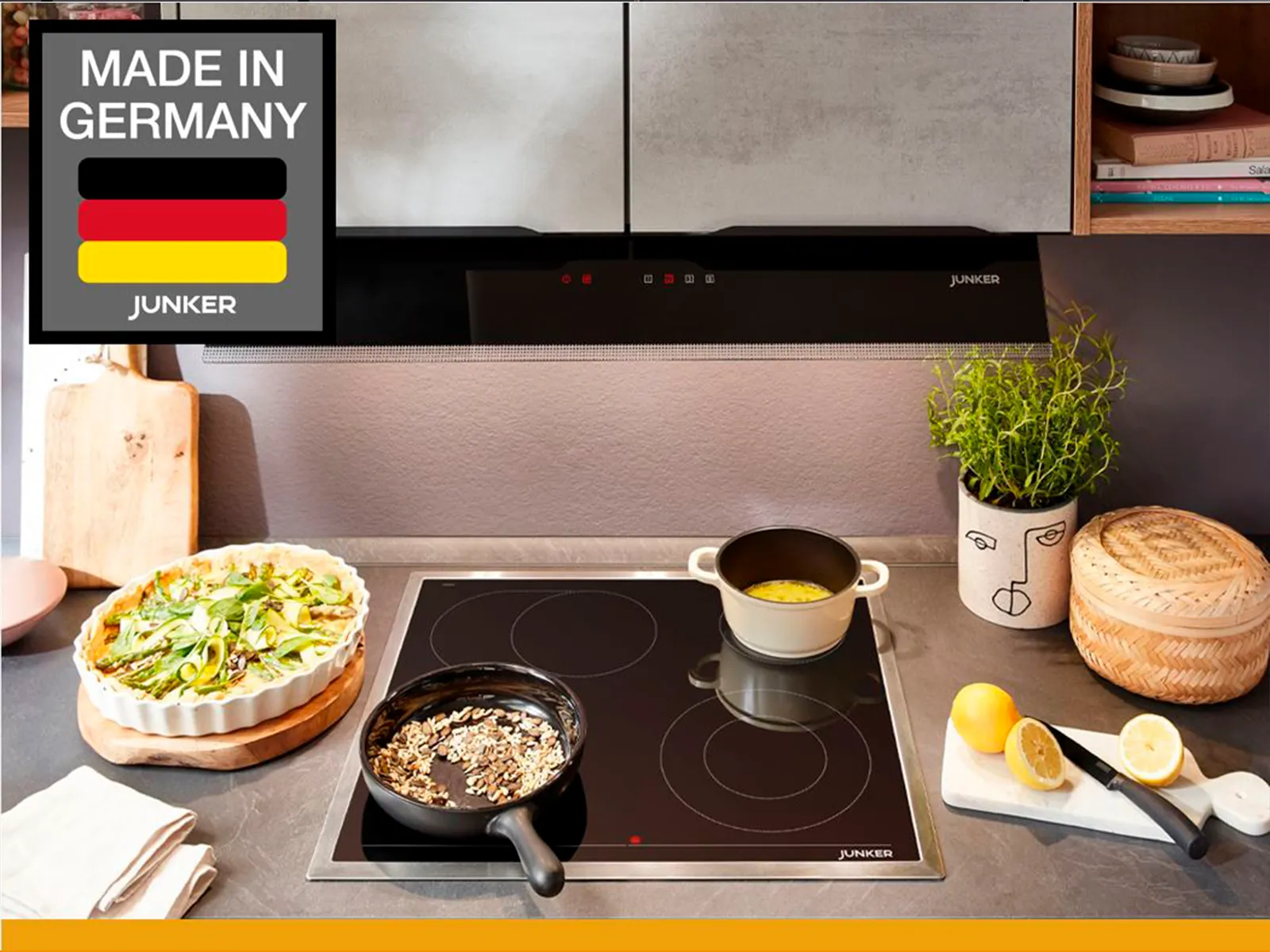 Moderne Küche mit einem Induktionskochfeld, Töpfen, einem Kuchen, frischen Kräutern und einem Schild mit der Aufschrift "Made in Germany" von Junker