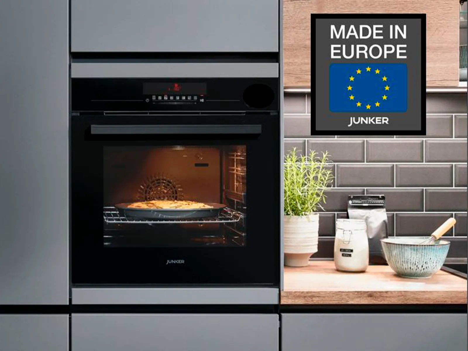 Moderne Küche mit schwarzem Ofen, „Made in Europe“-Schild, Pflanzen und Kochgeschirr auf der Arbeitsplatte. 