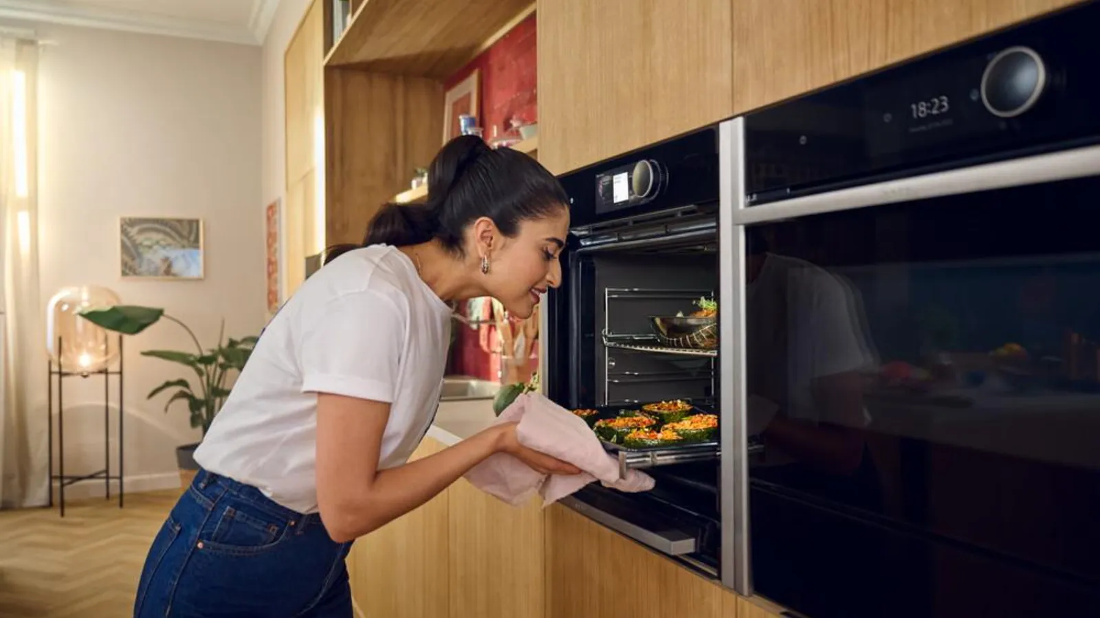 A woman with dark hair tied back, wearing a white t-shirt and jeans, smiles as she leans into a sleek, open black built-in oven. She holds a baking tray filled with stuffed vegetables or small dishes, using a light-colored cloth. The oven's digital display shows "18:23". To the right, another matching built-in oven is closed. The modern kitchen features light wooden cabinetry, a vibrant red tiled backsplash, and a countertop with a sink and fresh produce.