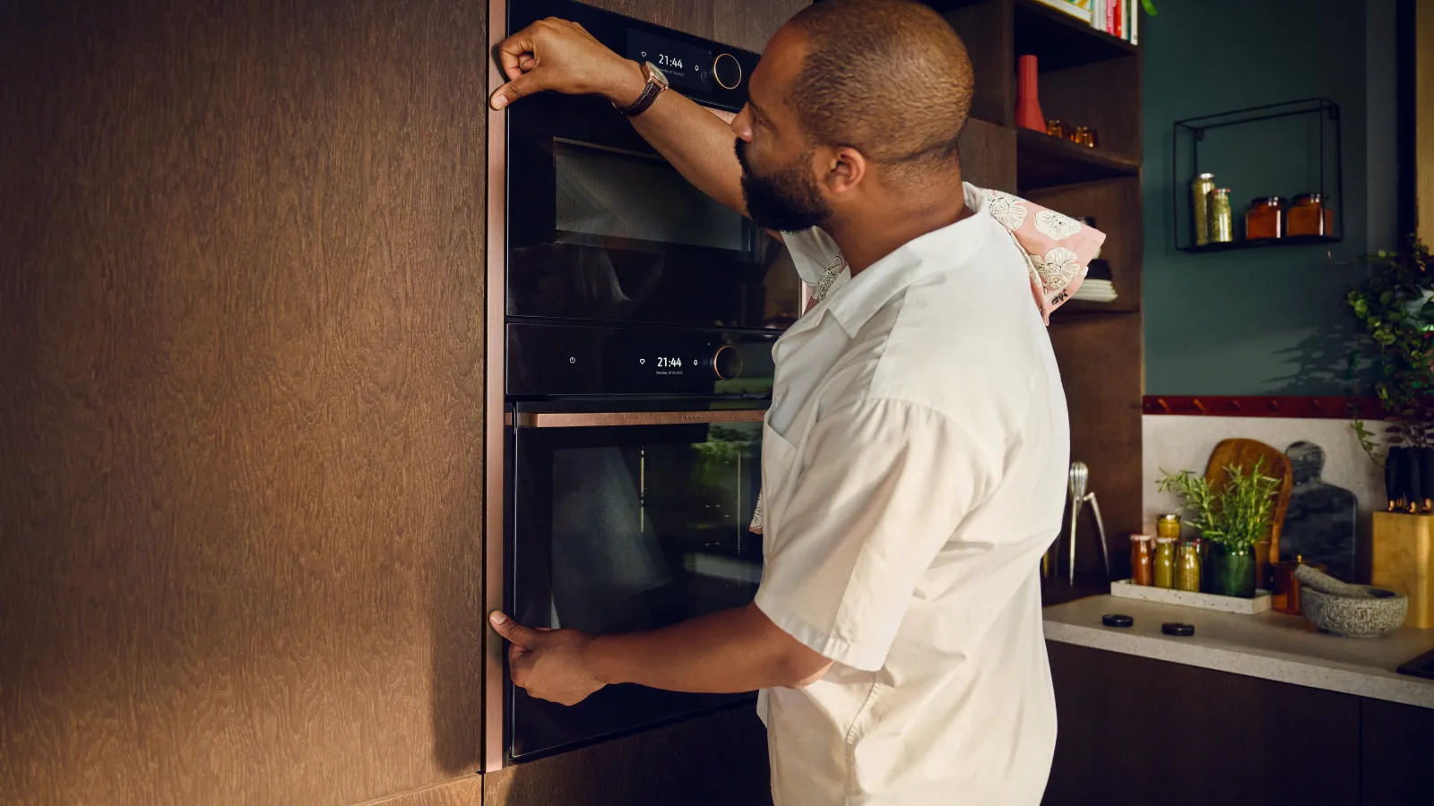 A man with a beard, wearing a white chef's coat, is adjusting the copper-colored trim around two sleek, black built-in ovens in a modern kitchen. The ovens are stacked vertically, with digital displays showing "21:44" and "HOME CONNECT". He has a pink and white patterned cloth draped over his shoulder. The ovens are integrated into dark brown, wood-grained cabinetry. To the right, the kitchen features a dark green wall with open wooden shelves holding cookbooks, jars of spices, and decorative items. A light grey countertop below is adorned with fresh herbs, cutting boards, a mortar and pestle, and a knife block.