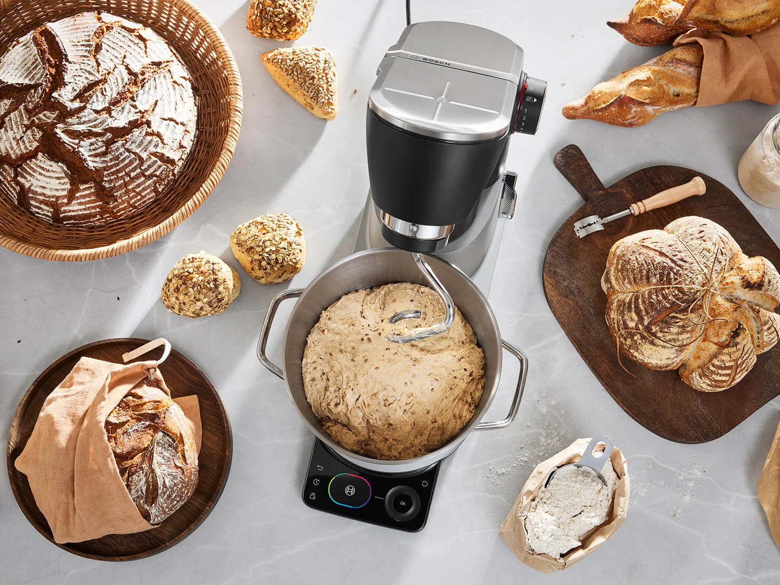 Bosch Series 8 stand mixer with kneading hook mixing bread dough, surrounded by artisan loaves, rolls, flour and baking tools on a light worktop.