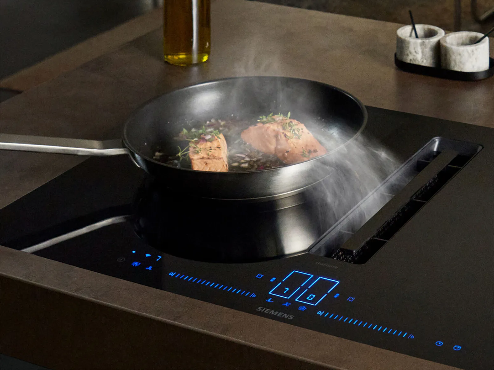 Salmon cooking in a pan on a Siemens induction hob with an active downdraft extractor, demonstrating fryingSensor technology.