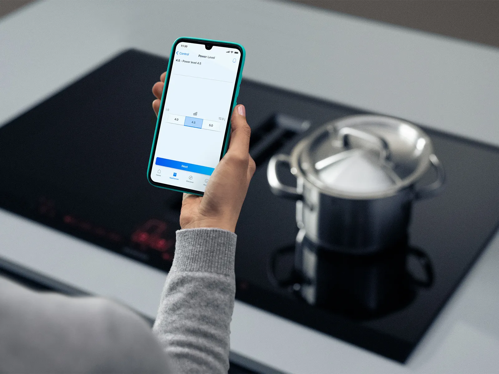 A hand holding a smartphone displaying a cooking app to control the power level of an induction hob with a pot on it.