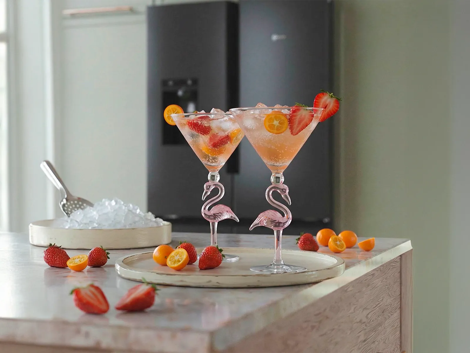 Two fruit-filled cocktails in flamingo-shaped glasses on a kitchen counter, with a NEFF MultiDoor refrigerator in the background.