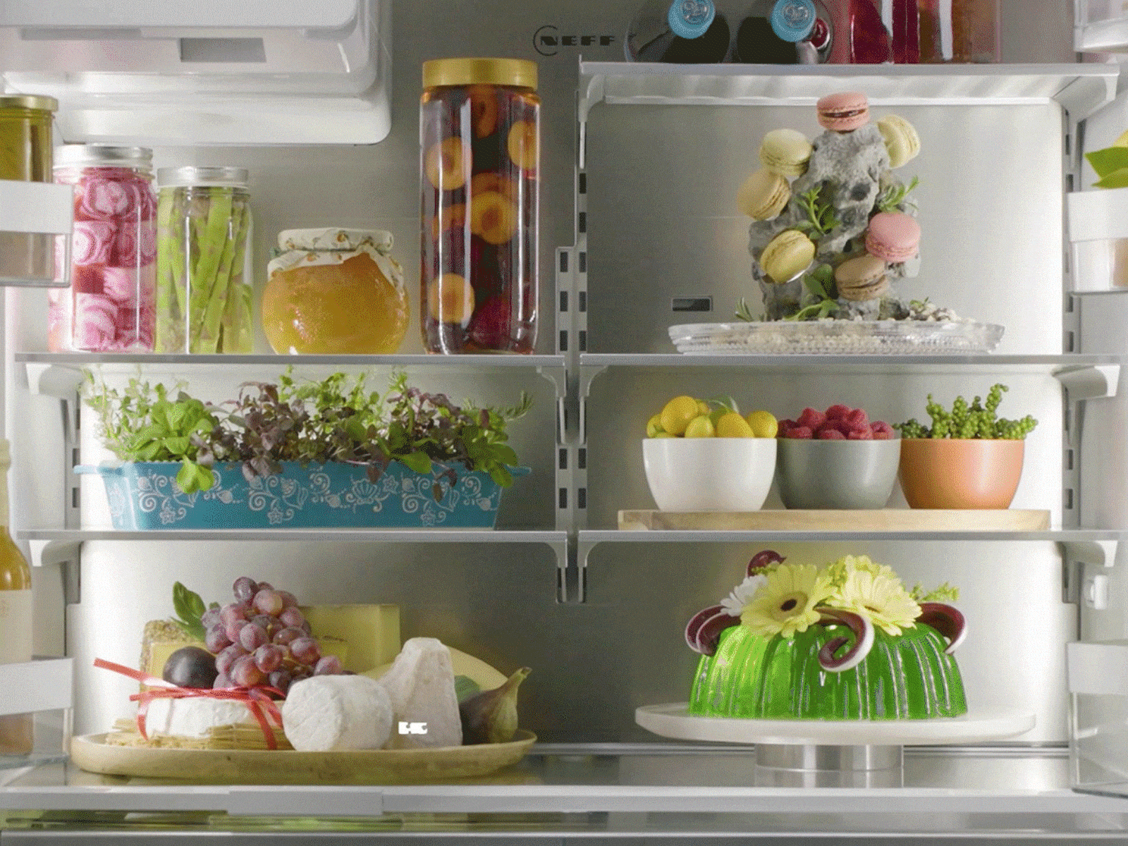 Inside an open NEFF MultiDoor refrigerator with neatly arranged jars, fresh herbs, fruit bowls, cheeses, and decorative desserts on glass shelves. Shelf setups and positions shuffle through.