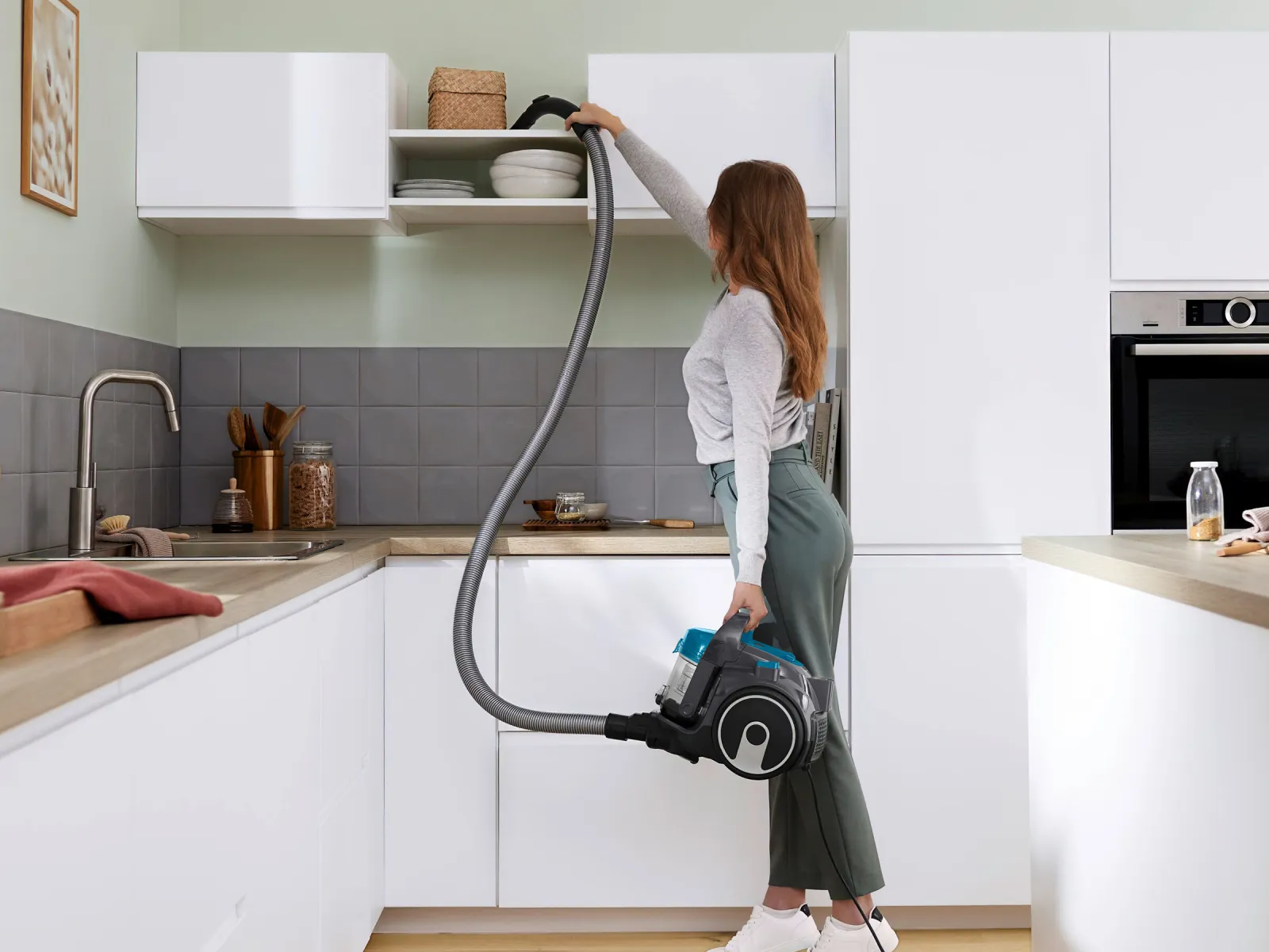 A woman vacuuming a kitchen shelf with a canister vacuum cleaner.