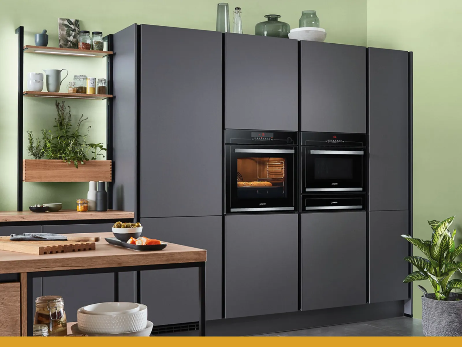 Cuisine moderne avec placards gris, fours intégrés, plan en bois, étagères avec plantes et ustensiles, et mur vert.