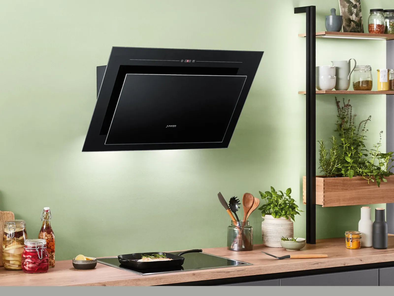 Cuisine moderne avec hotte noire murale au-dessus des plaques, entourée d'étagères en bois avec plantes et bocaux. 