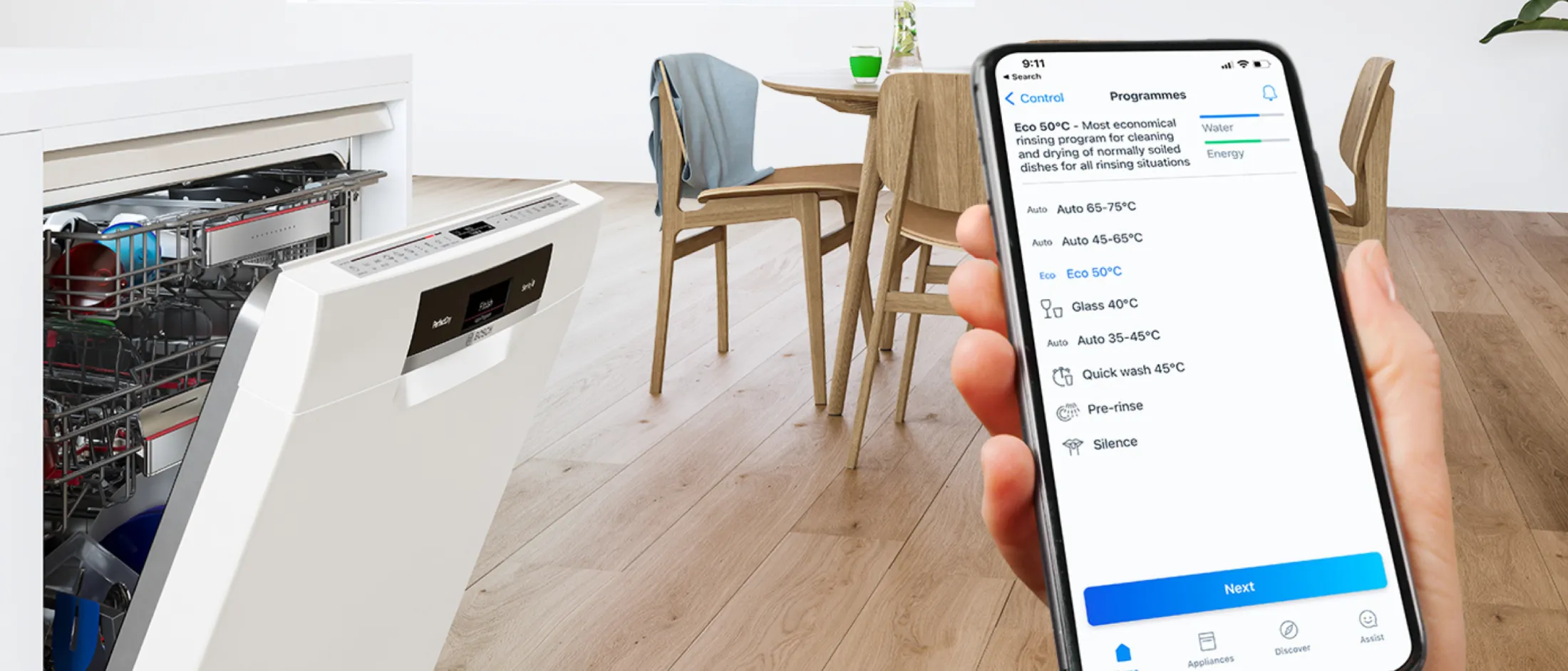 יד מחזיקה בטלפון חכם שמציג תוכניות לשימוש במדיח כלים. המדיח פתוח ומכיל כלים, ברקע כסאות מעץ וצמחייה.