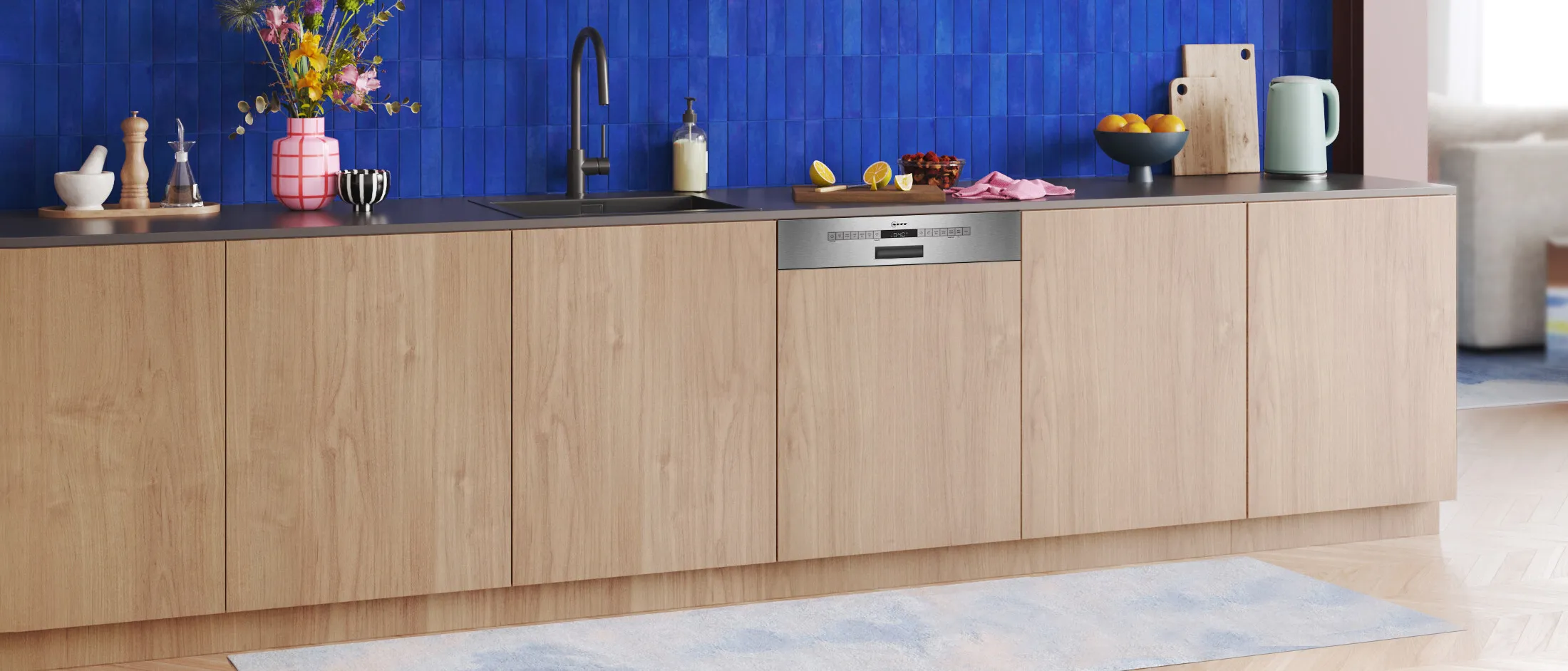 Modern kitchen with wooden cabinetry, a black countertop, blue tiled backsplash, and colorful decor, including flowers and fruit.