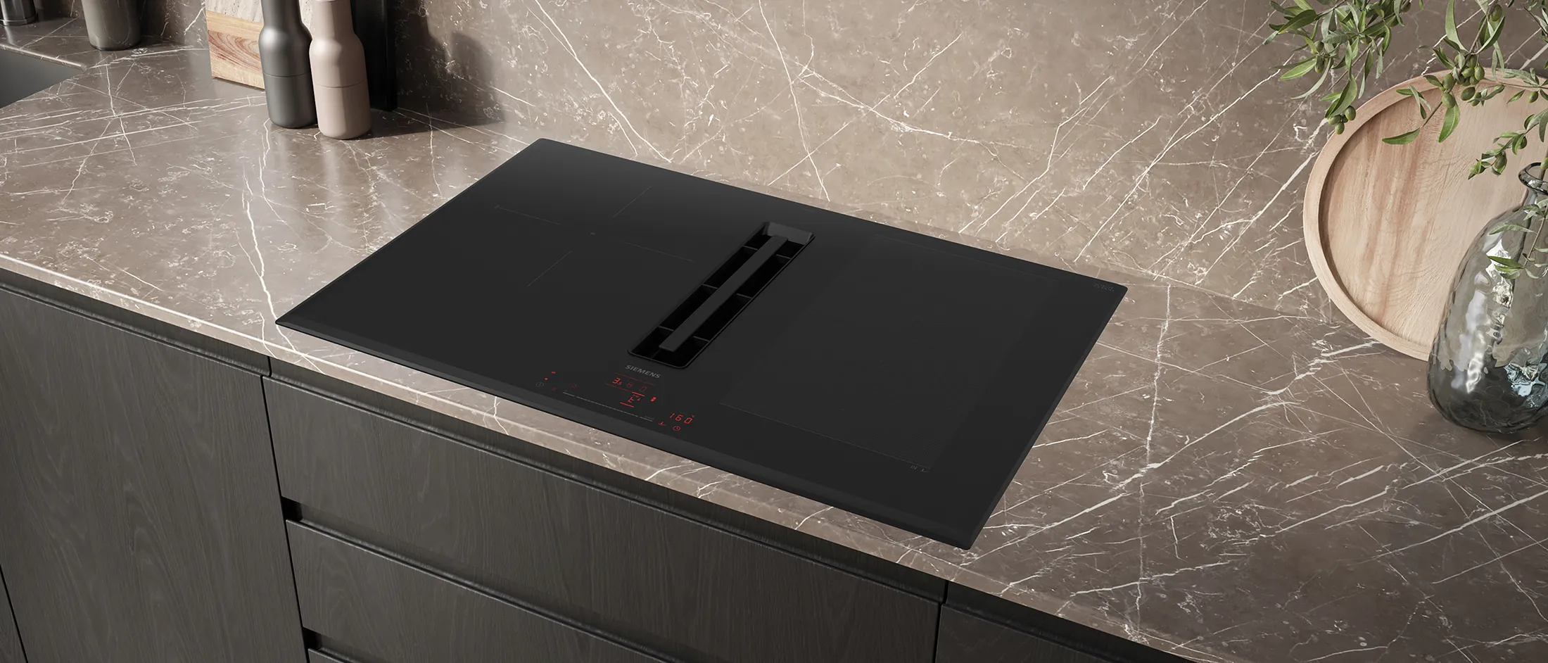 El extractor integrado en el centro de la placa de inducción Siemens absorbe el vapor emitido por una cacerola y donde se cuece una receta de verdura y una sartén en la que se cocinan dos raciones de salmón a las hierbas.