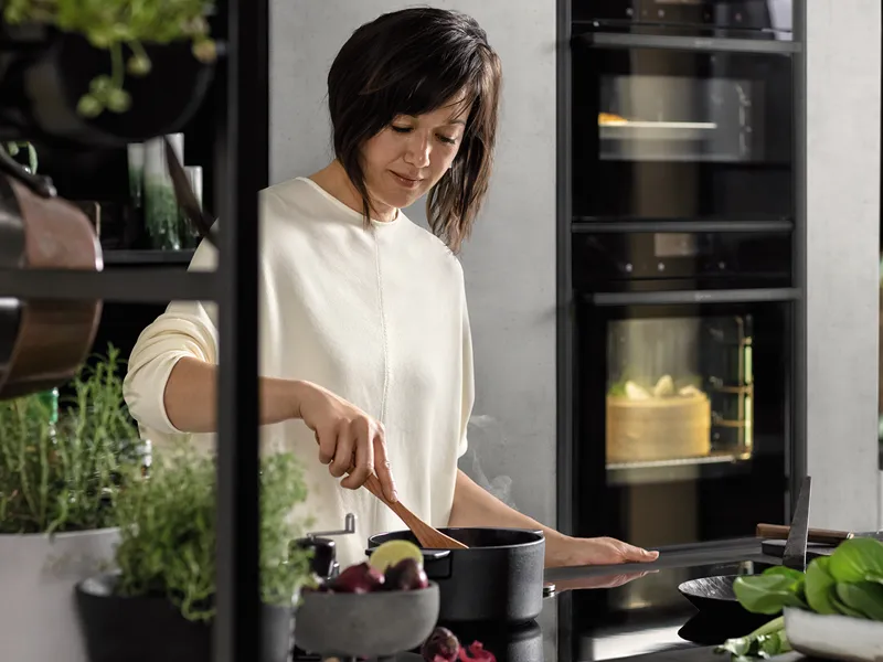 Γυναίκα με λευκό πουκάμισο μαγειρεύοντας στην κουζίνα με φυτά και συσκευές.