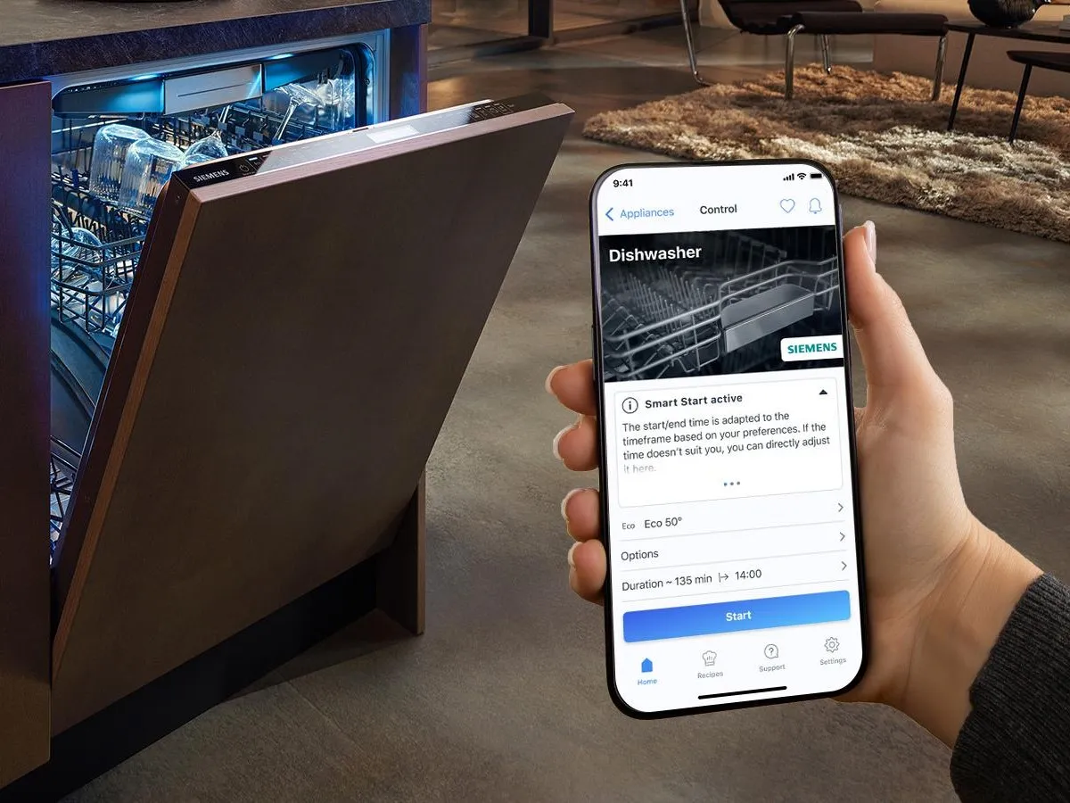 Una mano sostiene un móvil desde el que controla el lavavajillas que se encuentra en el fondo