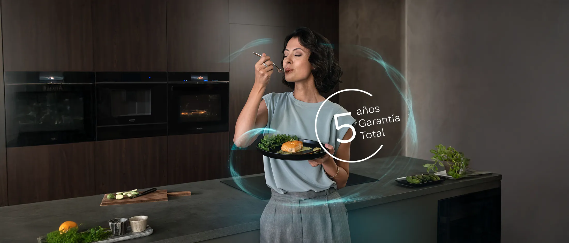 Una persona sostiene un plato negro con salmón y verduras en una cocina moderna, mostrando elegancia y una garantía total de cinco años.