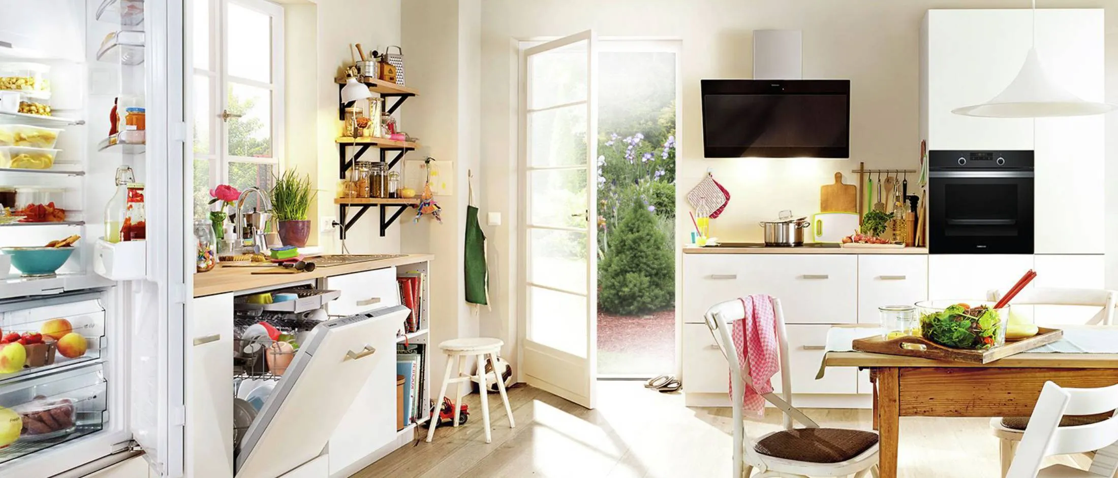 Lichte, moderne keuken met een goedgevulde koelkast, een houten eettafel en een open deur naar de tuin.