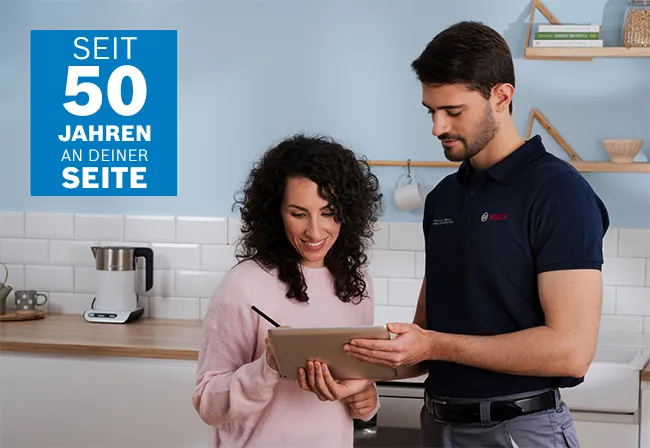 Das Bild zeigt einen Kücheninnenraum mit einem Mann, der eine Geschirrspülmaschine repariert, umgeben von modernen weißen Schränken und Geräten.