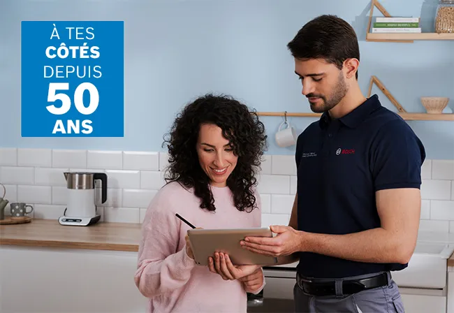 Un technicien Bosch en gants répare un appareil dans une cuisine moderne pendant qu'une femme observe attentivement. Des outils sont disposés sur le plan de travail à proximité.