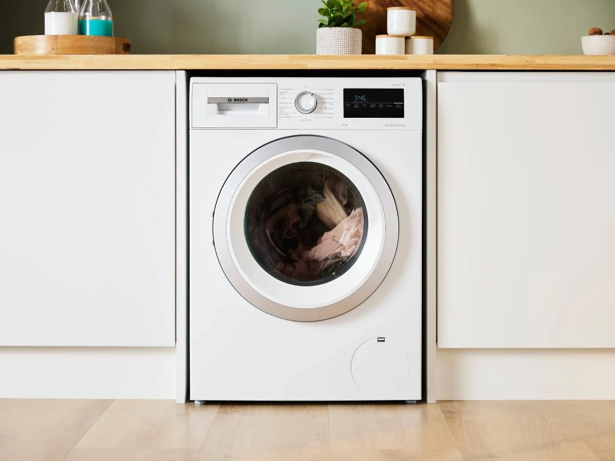 A white Bosch washing machine with clothes inside, integrated into a modern kitchen or laundry area with light wood countertops.