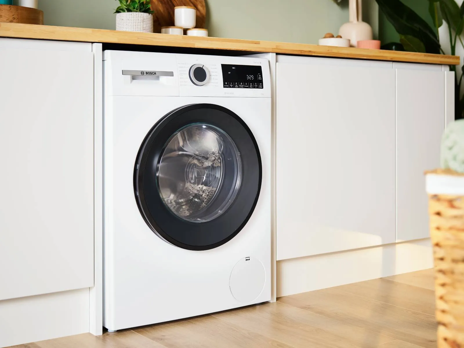 A white Bosch front-loading washing machine with a silver-ringed door, showing clothes inside, is installed under a light wood countertop between white cabinets. 